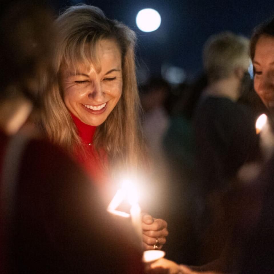 The image shows a woman smiling warmly while holding a lit candle during an evening event. She is surrounded by others who are also holding candles, creating a soft, glowing atmosphere. The background is dark, with the moon visible in the sky, suggesting a night-time vigil or celebration. The woman's joyful expression and the serene setting convey a sense of community and shared experience.