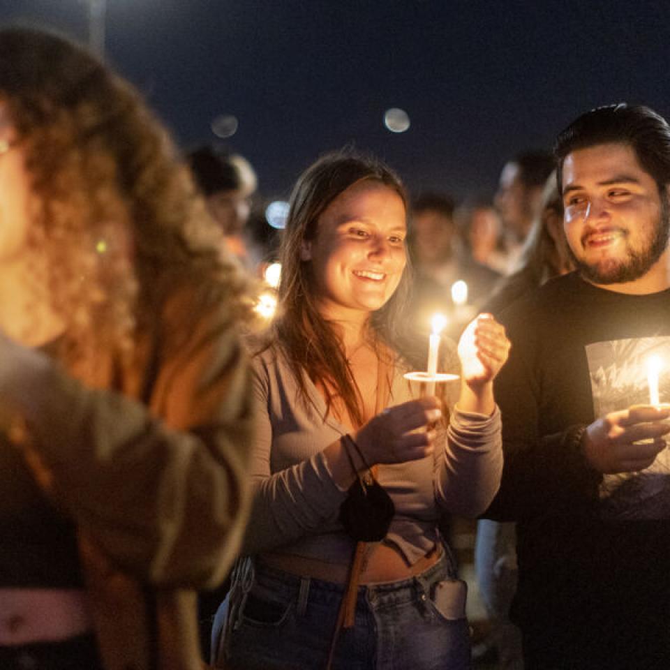 The image depicts a group of people gathered outdoors at night, participating in a candlelight event. The focus is on three individuals in the foreground, smiling and holding lit candles protected by small cups. The warm glow of the candles illuminates their faces, creating a serene and communal atmosphere. The background is filled with more people holding candles, all contributing to a peaceful and reflective scene.