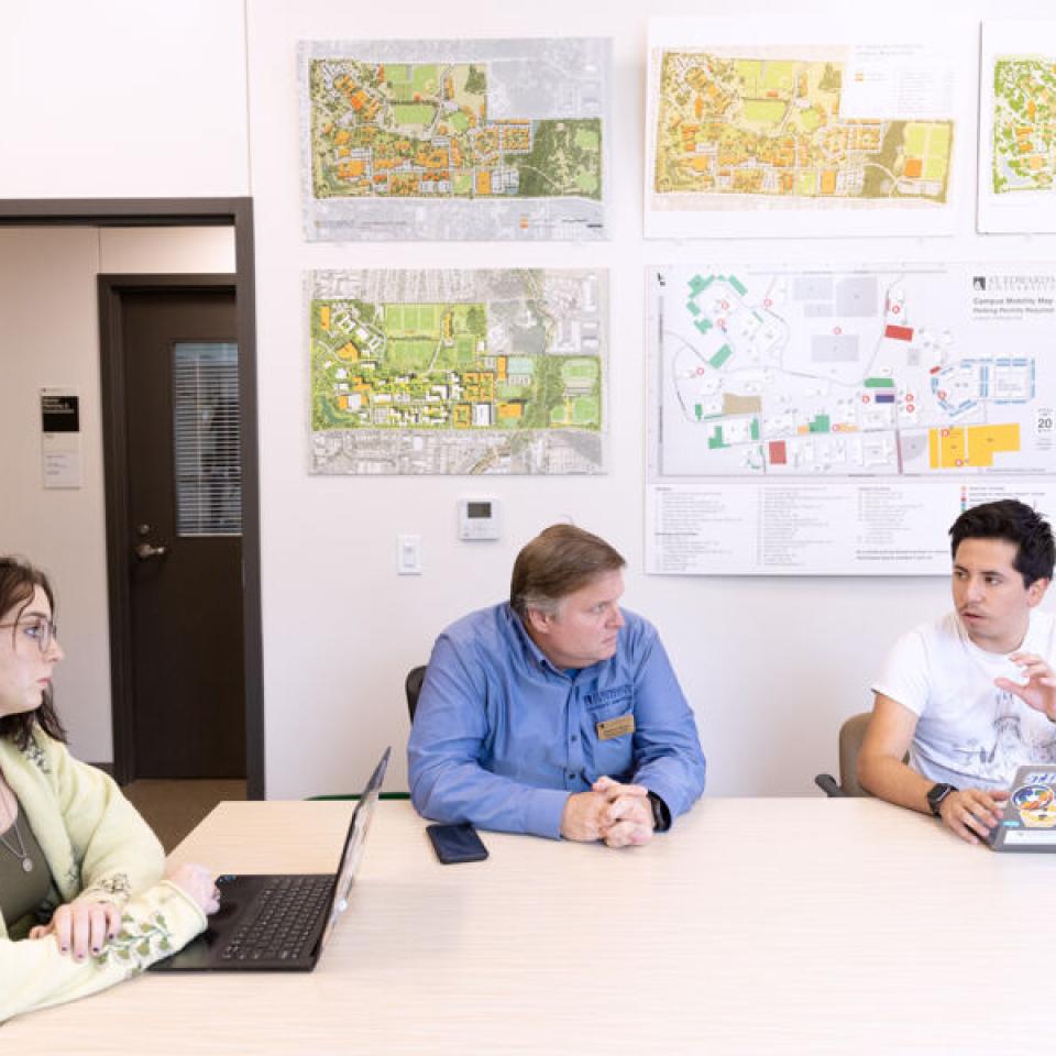 Ethan Tobias, right, speaks with Jim Morris, middle, and another sustainability intern as they sit at a conference room with laptops on the table and maps on the wall behind them.