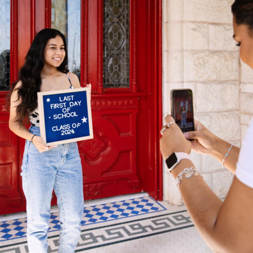 student posing with first day of class sign by red doors