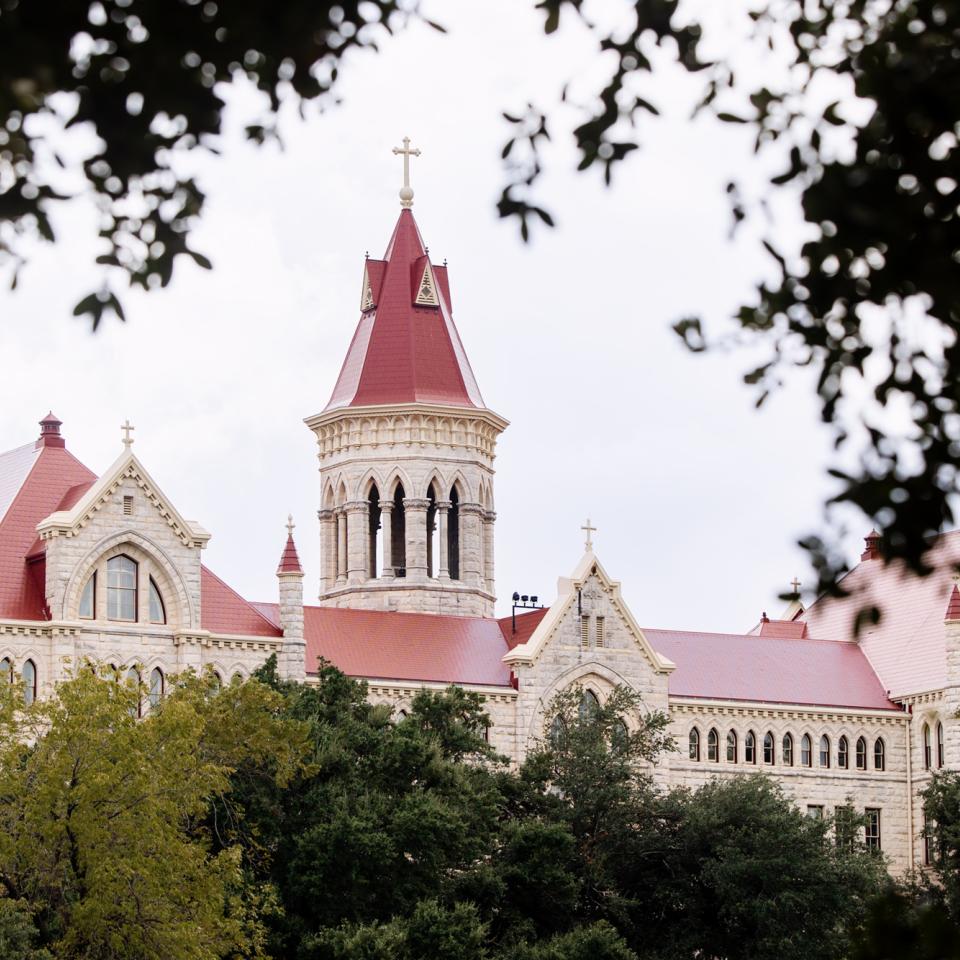 Main Building is seen with its distinctive red roofs and a central tower, surrounded by lush greenery. The building is framed by dark foliage in the foreground, creating a picturesque view. The architecture features pointed gables and ornate detailing, typical of a classic academic institution. The sky appears overcast, adding a soft light to the scene, which emphasizes the building's prominence and elegance.