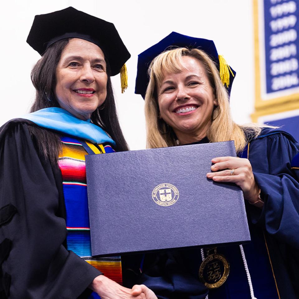 The image shows two women in academic regalia during a commencement ceremony. The woman on the left is wearing a black cap and gown with a colorful stole, while the woman on the right is in a dark blue gown with gold accents, holding a diploma folder. Both are smiling and looking at the camera. The background features a banner with a list of years, suggesting the setting is a university or college gymnasium during a graduation event.