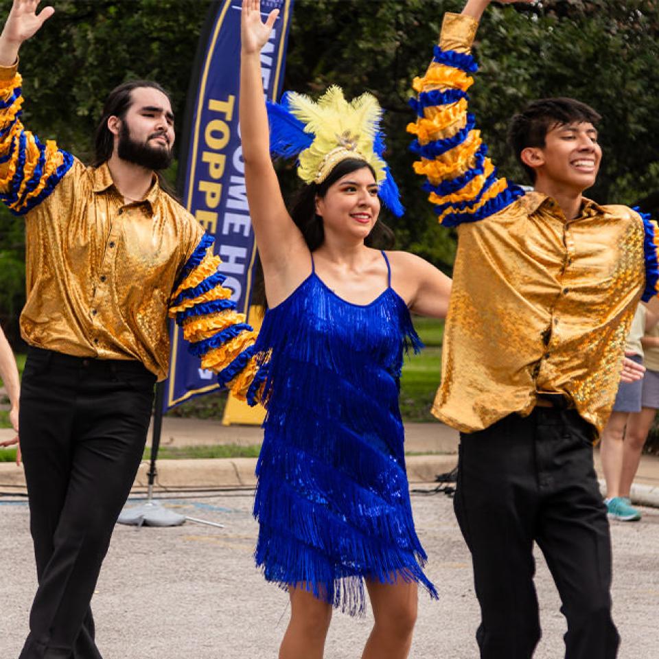 The image captures a vibrant outdoor performance featuring dancers in colorful costumes. Two men in shiny gold shirts and black pants and two women in blue fringe dresses with yellow and blue feathered headpieces are dancing with their arms raised. They are performing in front of a "Homecoming" banner, indicating a festive event. The dancers are smiling and appear to be enjoying the performance, while people in the background watch, adding to the lively atmosphere.