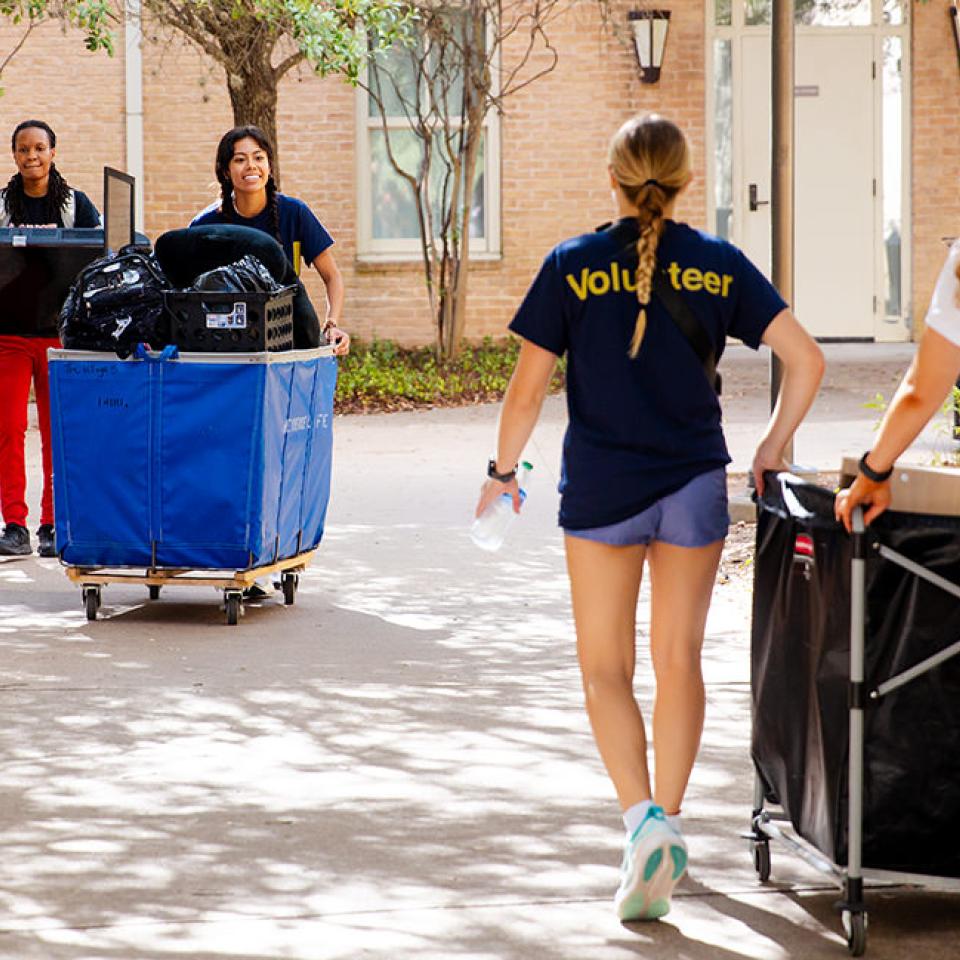 The image shows several young people moving items on a sunny day. A young man in a white T-shirt and black shorts walks while holding a piece of paper. Behind him, two volunteers in navy blue shirts, labeled "Volunteer," push large carts filled with bags and belongings. Another young man in red pants carries a flat-screen TV. They are walking on a path surrounded by trees and a brick building. The atmosphere is energetic and collaborative, suggesting a group effort in moving or setting up an event.