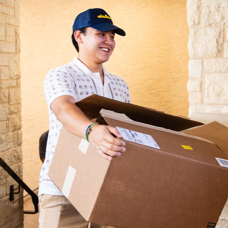 The image shows a young man smiling while carrying a large cardboard box. He is wearing a white patterned polo shirt, beige pants, and a navy blue cap. The setting appears to be outside, near a building entrance made of stone. The atmosphere is bright and cheerful, indicating a positive and possibly exciting occasion, such as moving in or helping with an event. The young man seems happy and engaged in his task.