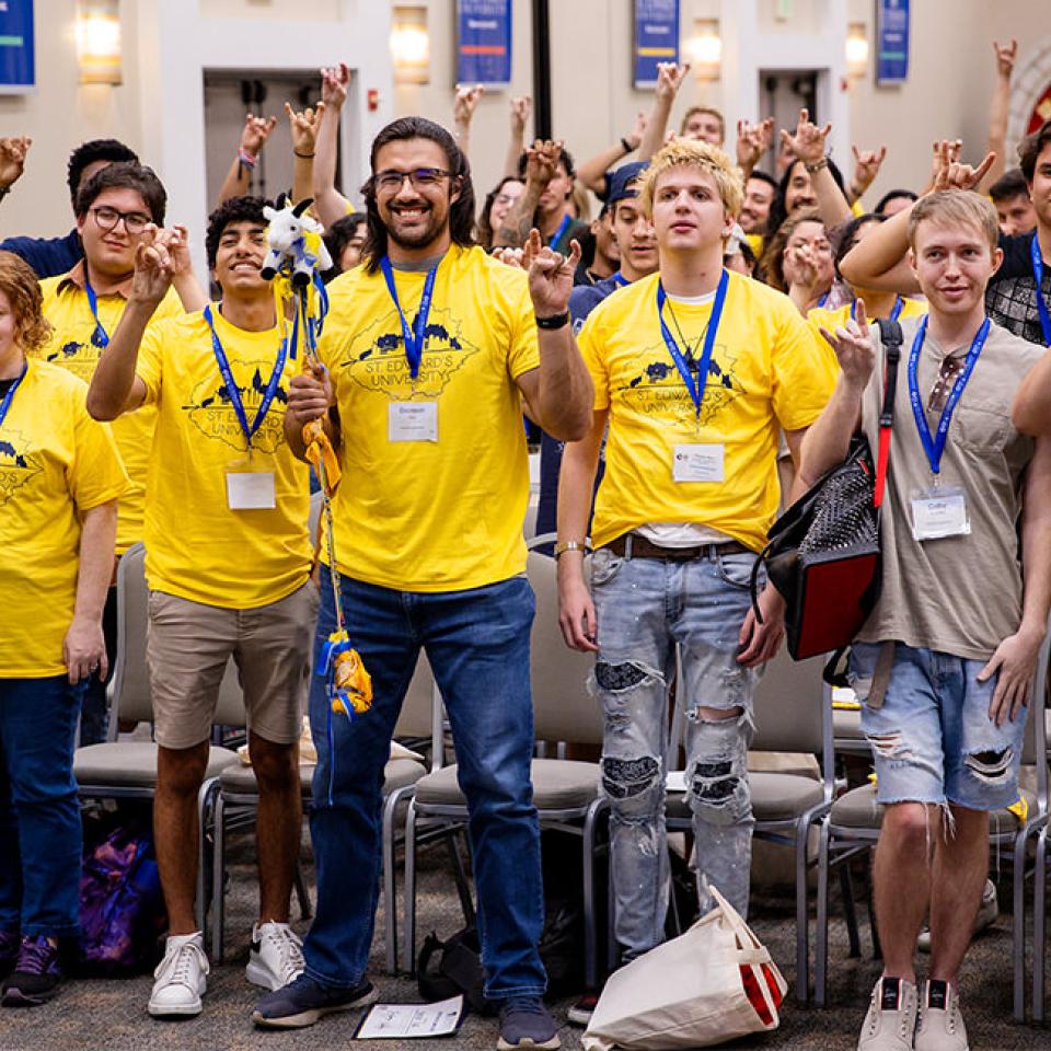 The image shows a large group of enthusiastic students standing together, all wearing bright yellow T-shirts with "St. Edward's University" printed on them. They are making hand gestures, likely a school spirit sign, and many are wearing name tags and lanyards. The setting appears to be a spacious, well-lit hall with chairs arranged in rows. The atmosphere is lively and energetic, with everyone smiling and participating in the group activity or event.