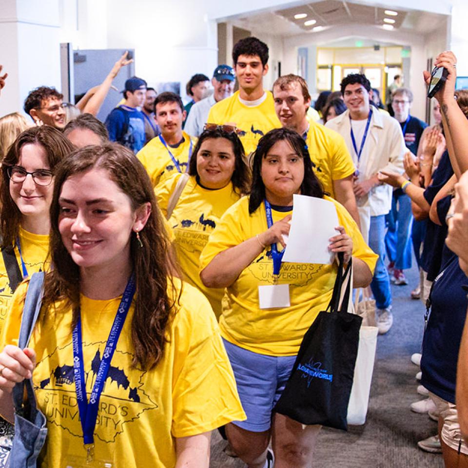 The image shows a group of students wearing bright yellow T-shirts with "St. Edward's University" printed on them, walking through a hallway. They are being welcomed and cheered on by people lining both sides, clapping and raising their hands. The atmosphere is celebratory and energetic, with the students smiling and looking excited. Many students are wearing lanyards and name tags, and some are carrying bags or papers. The hallway is well-lit and bustling with activity.