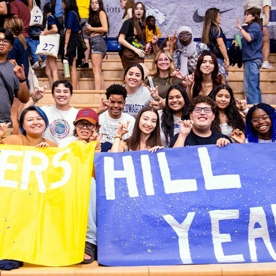 The image shows a group of excited students sitting in bleachers at a pep rally, holding up two large banners. The yellow banner reads "Toppers Up," and the blue banner says "Hill Yeah." The students are smiling, making hand signs, and showing their school spirit. The background is filled with more students and some activity, suggesting a lively and energetic atmosphere. The diverse group is dressed in casual attire, and their expressions reflect enthusiasm and camaraderie.