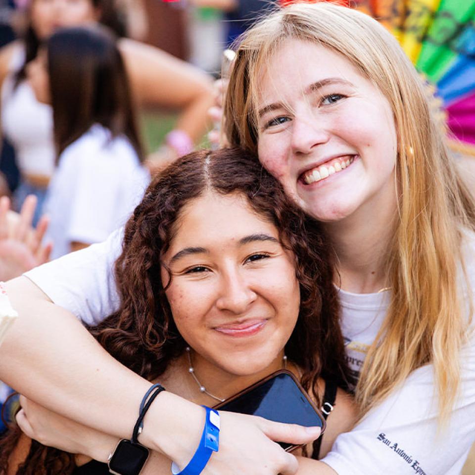 The image captures a joyful moment between two young women at a block party. They are smiling broadly and hugging each other. One woman is holding an ice cream bar, while the other has a phone in her hand. In the background, other party attendees are visible, adding to the lively and festive atmosphere. The setting appears to be outdoors with colorful elements like a rainbow wheel, suggesting a fun and vibrant community event.