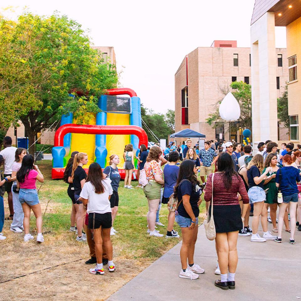 The image shows a lively outdoor block party with a large crowd of people socializing and enjoying various activities. In the center, there is a colorful inflatable slide, and around it, groups of people are talking and mingling. The event is set in a grassy area surrounded by buildings, with trees providing shade. Attendees are dressed in casual summer attire, and the atmosphere is vibrant and cheerful, suggesting a fun and engaging community event.