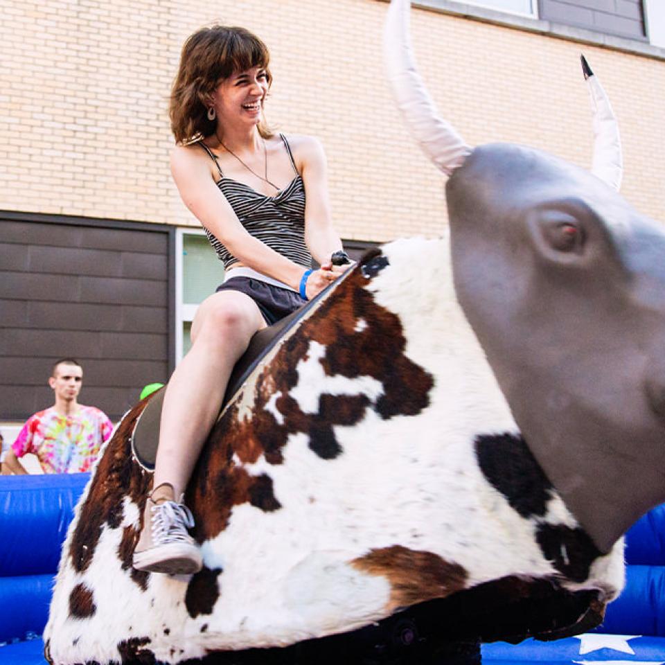 The image shows a young woman laughing and enjoying herself while riding a mechanical bull at an outdoor event. She is wearing a striped tank top and shorts. The mechanical bull is set up in an inflatable ring with blue padding. In the background, several onlookers, including a person in a tie-dye shirt and others in casual attire, are watching the activity. The setting appears to be a festive block party, with the building and outdoor decorations adding to the lively atmosphere.