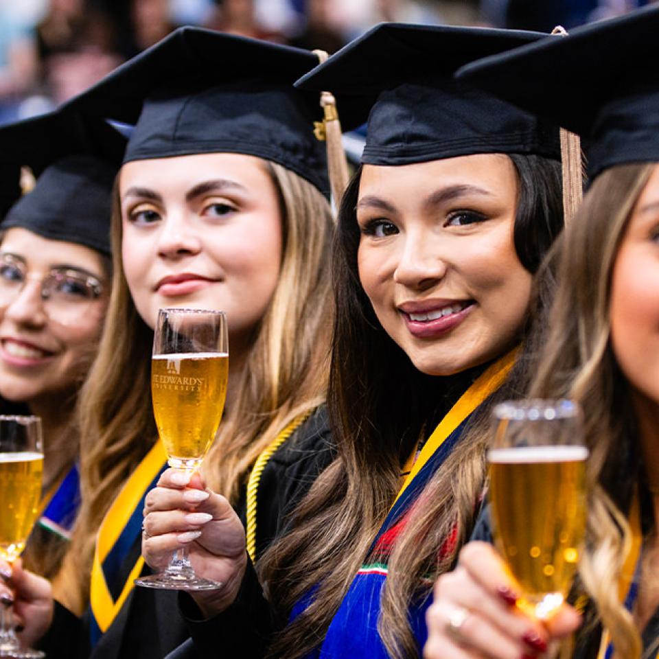 The image features a row of female graduates wearing black caps and gowns, sitting side by side. They are smiling and holding up champagne glasses, celebrating their accomplishment. Each glass is etched with the logo of St. Edward's University. The graduates are adorned with various honor cords and stoles, signifying their academic achievements. The background is blurred, focusing on the joyful expressions of the graduates as they look towards the camera.