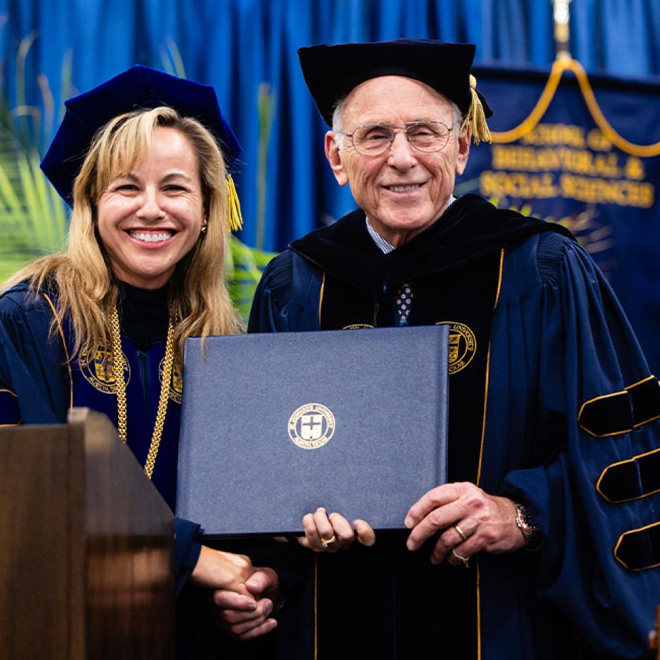 The image shows two academic figures, a man and a woman, dressed in formal graduation regalia, standing together and smiling. The woman, on the left, has long blonde hair and wears a navy blue gown with gold accents. The man, on the right, has gray hair, glasses, and wears a black cap and gown with gold trim. He is holding a diploma or certificate. They are standing in front of a podium with the university emblem and a backdrop featuring blue and gold banners, indicating a formal commencement ceremony.