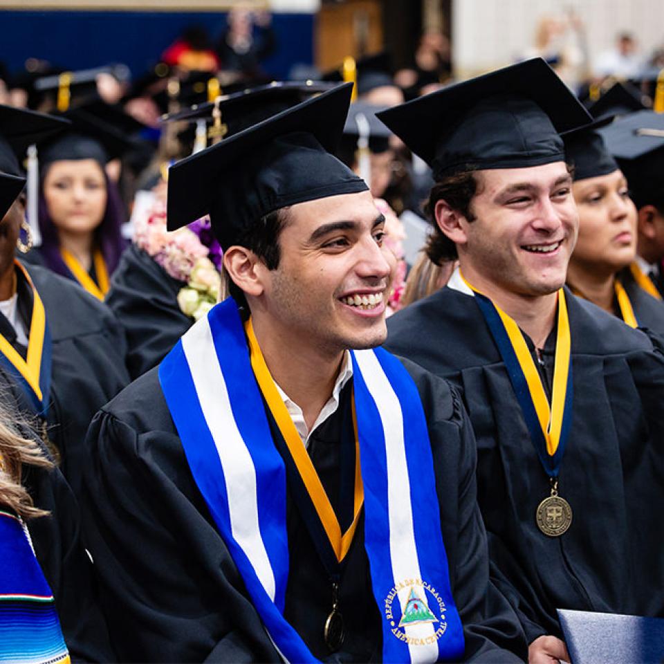 The image shows a group of graduates seated at their commencement ceremony, smiling and looking ahead. They are wearing black caps and gowns, with some adorned with colorful stoles and medallions. The atmosphere is joyous and celebratory, as the graduates are clearly happy and proud of their accomplishments. The background is filled with more graduates and the setting appears formal, indicating a well-organized and significant event.