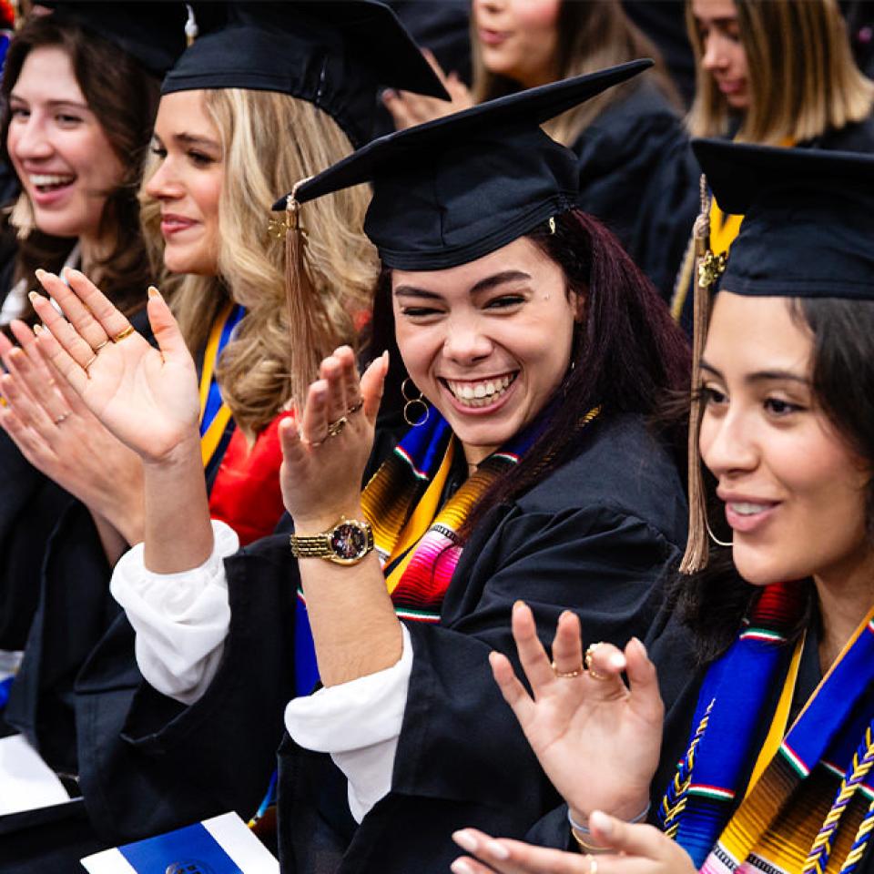 The image shows a group of graduates sitting in their seats during a commencement ceremony, clapping and smiling with joy. They are wearing black caps and gowns, some adorned with colorful stoles. The focus is on a woman in the center who is beaming with happiness. The graduates around her are equally joyful, celebrating their achievement. The atmosphere is festive and filled with excitement, reflecting the pride and accomplishment of the graduates.