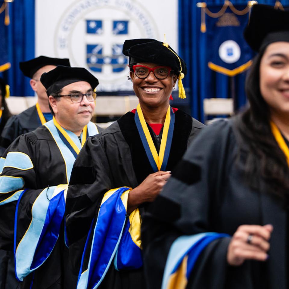 The image shows a group of graduates in academic regalia, processing during a commencement ceremony. The focus is on a smiling graduate wearing a black gown, cap, and red glasses, with a gold medallion around the neck. Other graduates, also in black gowns with blue and gold accents, follow in line. The background features the St. Edward's University emblem and blue and gold decorations, creating a formal and celebratory atmosphere. The scene reflects pride and achievement among the graduates.