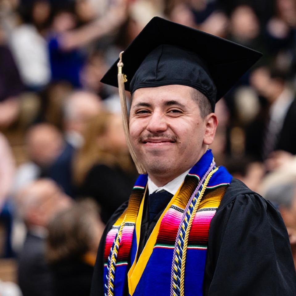 The image shows a proud graduate at a commencement ceremony, smiling broadly. He is wearing a black graduation cap and gown, with a colorful serape stole and a gold medallion. The background is filled with blurred spectators, suggesting a large and supportive audience. The graduate's expression reflects joy and accomplishment, capturing the celebratory atmosphere of the event.