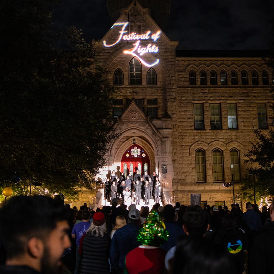 The image shows a nighttime event called the "Festival of Lights" at St. Edward's University. A crowd is gathered in front of a beautifully lit stone building, watching a choir perform on the steps. The building has "Festival of Lights" projected onto it, adding to the festive atmosphere. The crowd is attentive, with some people wearing holiday-themed attire, including a Christmas tree hat. The scene is warm and inviting, highlighting the communal and celebratory spirit of the event.