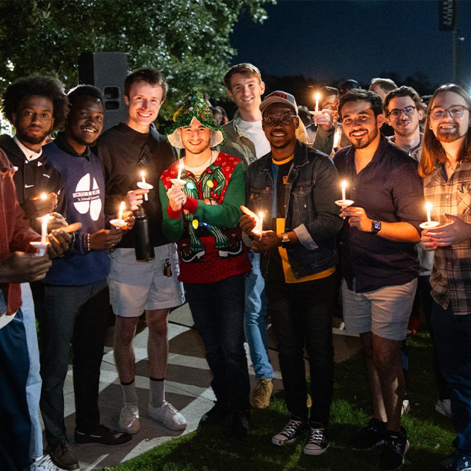 The image shows a group of friends at a candlelight vigil, standing close together and smiling. Each person holds a lit candle, and the group is dressed in casual attire, with one person wearing a festive holiday sweater and hat. The setting is outdoors at night, with trees and other participants visible in the background. The atmosphere is warm and communal, reflecting the spirit of unity and celebration at the event. The city skyline is faintly visible in the distance, adding to the serene ambiance.