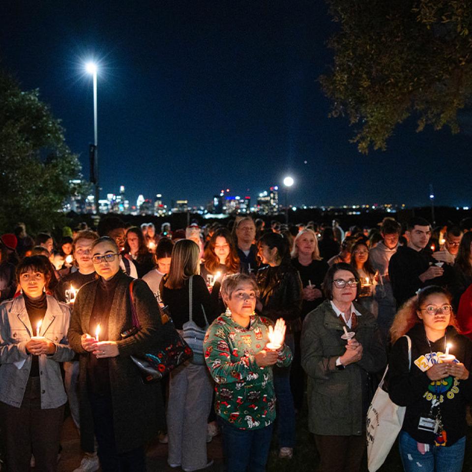 The image shows a large group of people gathered outdoors at night for a candlelight vigil. Each person holds a lit candle, creating a warm glow against the dark sky. The crowd is diverse, with attendees of different ages and backgrounds, some wearing jackets and sweaters. In the background, city lights are visible, enhancing the serene and contemplative atmosphere of the event. The gathering appears solemn and reflective, indicating a moment of remembrance or solidarity.