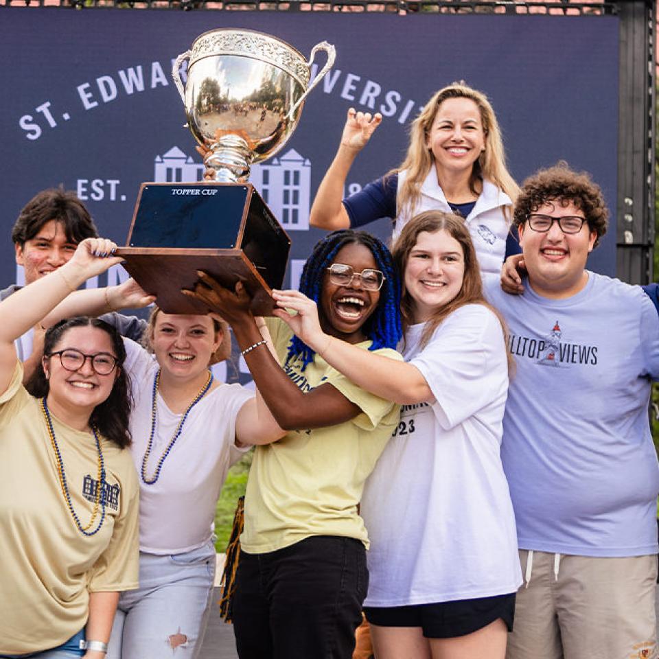 The image shows a group of happy students celebrating with a large trophy labeled "Topper Cup" at a St. Edward's University event. They are posing in front of a backdrop with the university's name and logo. The students are smiling broadly, and some are wearing beaded necklaces and university T-shirts. The atmosphere is festive and joyful, reflecting their excitement and pride in winning the trophy. The diverse group stands together, showcasing camaraderie and school spirit.