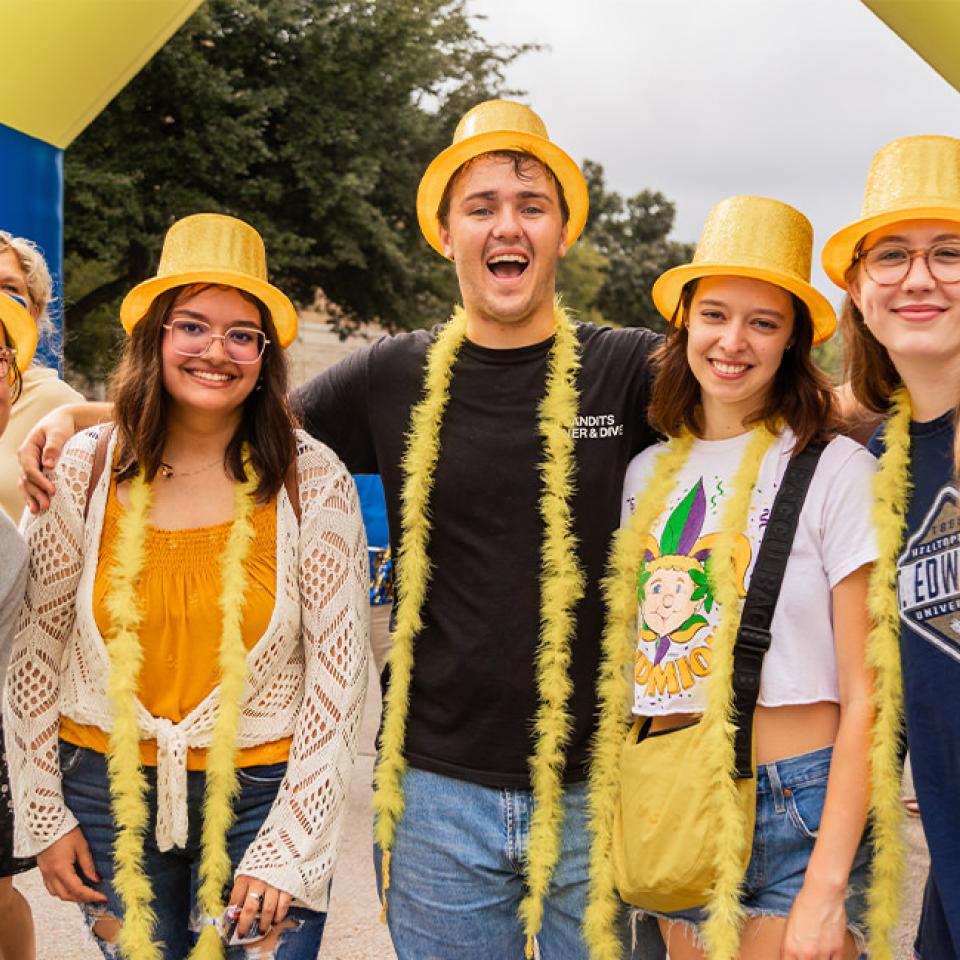 The image shows a group of five friends posing happily at an outdoor event. They are all wearing bright yellow hats and feather boas, adding a festive touch to their casual attire. One person is giving a peace sign, while the others smile broadly. They are standing under an inflatable arch with "SEU Hilltoppers" branding, indicating a school event or homecoming celebration. The atmosphere is cheerful and fun, with trees and other participants visible in the background.