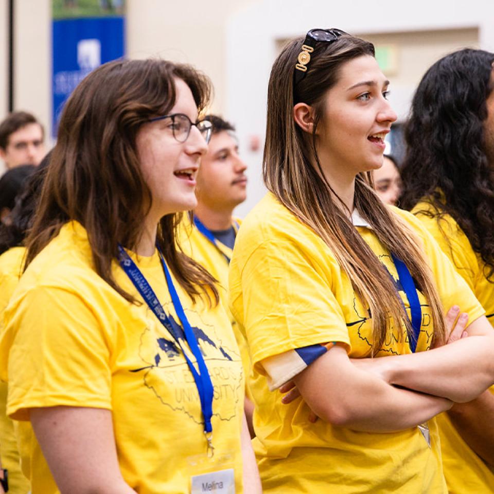 The image shows a group of young adults standing and smiling, wearing bright yellow T-shirts with "St. Edward's University" printed on them. They are participating in what appears to be an orientation or group activity, as many are wearing name tags and lanyards. The atmosphere is lively and positive, with everyone looking engaged and enthusiastic. In the background, there are blue banners and a well-lit interior, suggesting a formal event setting.