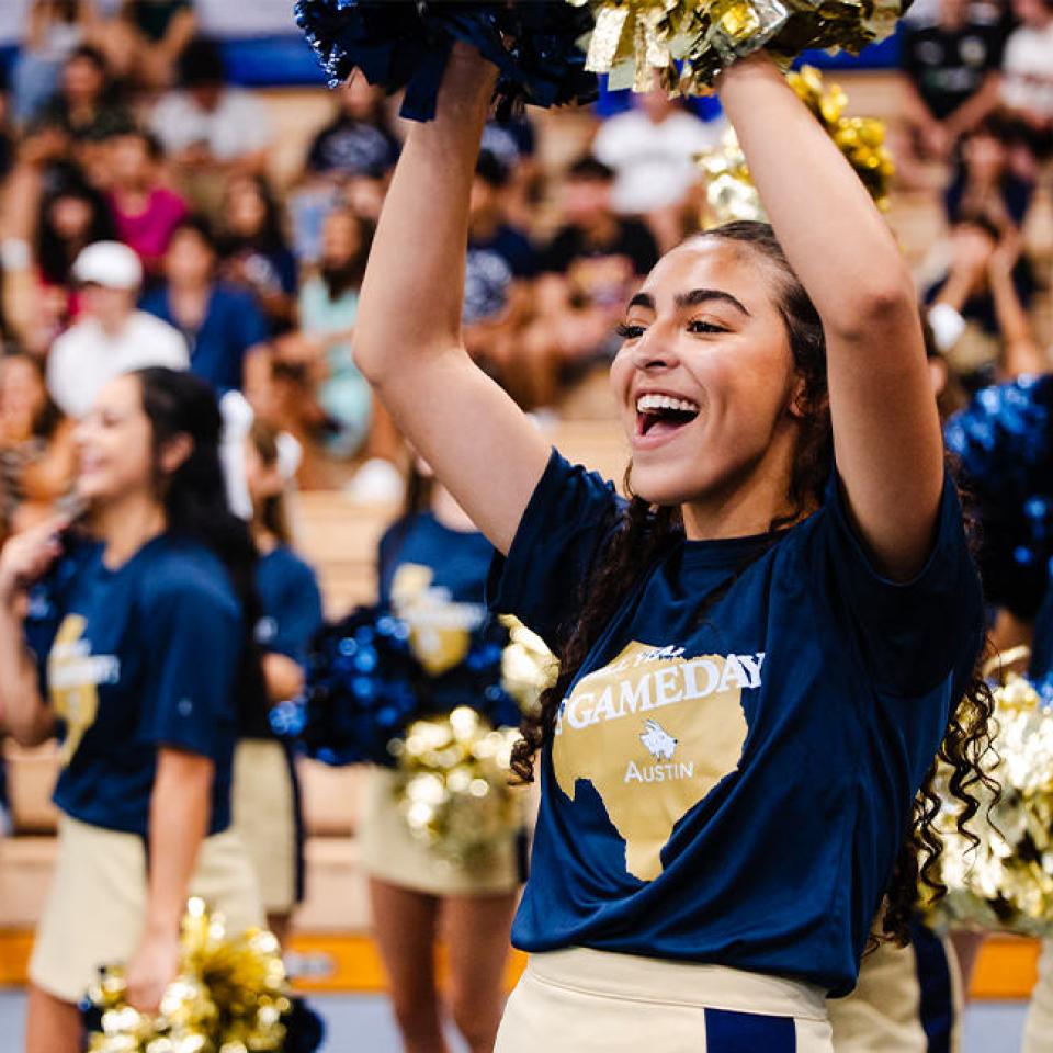 The image shows a group of enthusiastic cheerleaders performing at a pep rally. The focus is on a smiling cheerleader in the foreground, wearing a navy blue shirt with "Gameday Austin" printed on it and holding gold and blue pom-poms. She is raising her arms, cheering energetically. Other cheerleaders in similar outfits are visible in the background, along with a crowd of spectators in the bleachers, creating a lively and spirited atmosphere. The scene exudes excitement and school spirit.