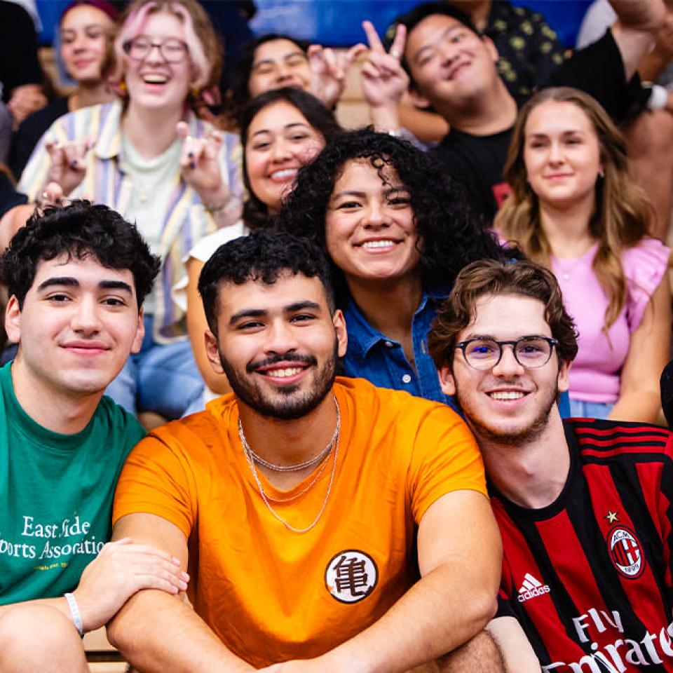 The image shows a group of enthusiastic young people sitting together in bleachers at a pep rally. In the front row, five friends are smiling brightly, with two of them making peace signs. The crowd behind them is equally animated, with students making various hand gestures and cheering. Everyone looks happy and engaged, creating a lively and spirited atmosphere. The diverse attire, including sports jerseys and casual wear, reflects a casual and friendly gathering.