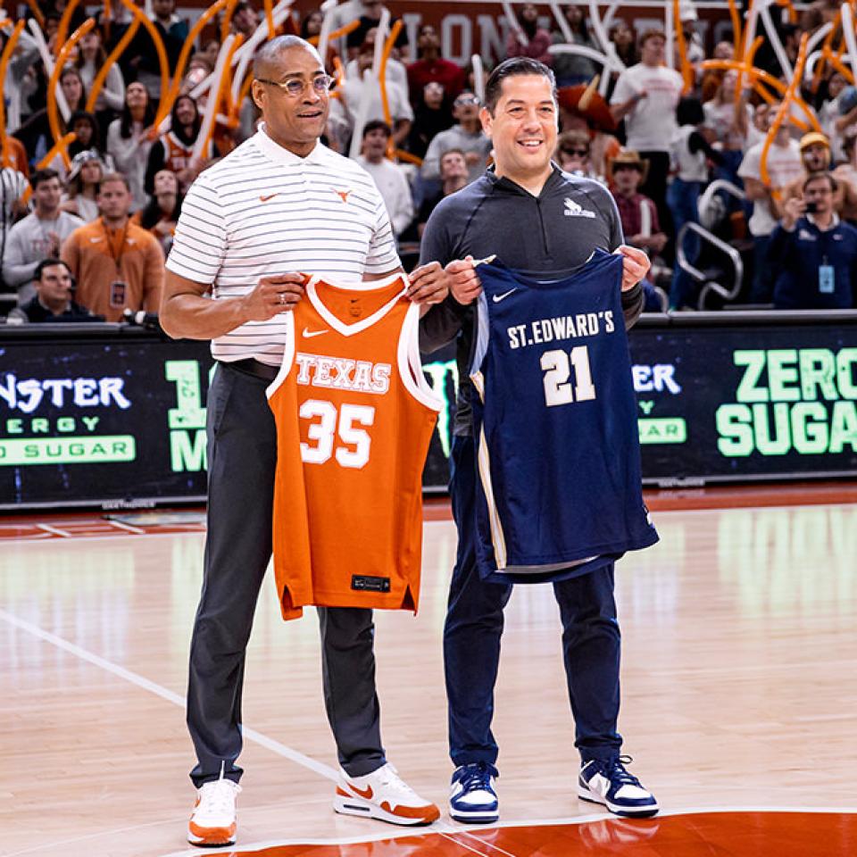 The image shows two men standing on a basketball court, each holding a jersey. The man on the left is holding an orange Texas jersey with the number 35, while the man on the right holds a navy blue St. Edward's jersey with the number 21. They are smiling and standing side by side, with a crowd of cheering fans and waving orange inflatable sticks in the background. The setting is a high-energy basketball game, highlighted by the lively crowd and the visible branding on the court.