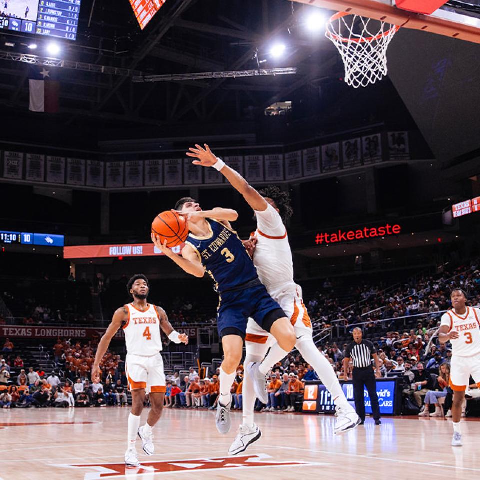 The image captures an intense moment in a basketball game between St. Edward's University and the Texas Longhorns. A player from St. Edward's (wearing a navy jersey, number 3) is driving towards the basket while being closely defended by a Texas player. Other players from both teams are visible on the court, and the crowd watches intently. The scoreboard shows the score and time remaining, and the arena is well-lit with banners and digital displays adding to the dynamic atmosphere.