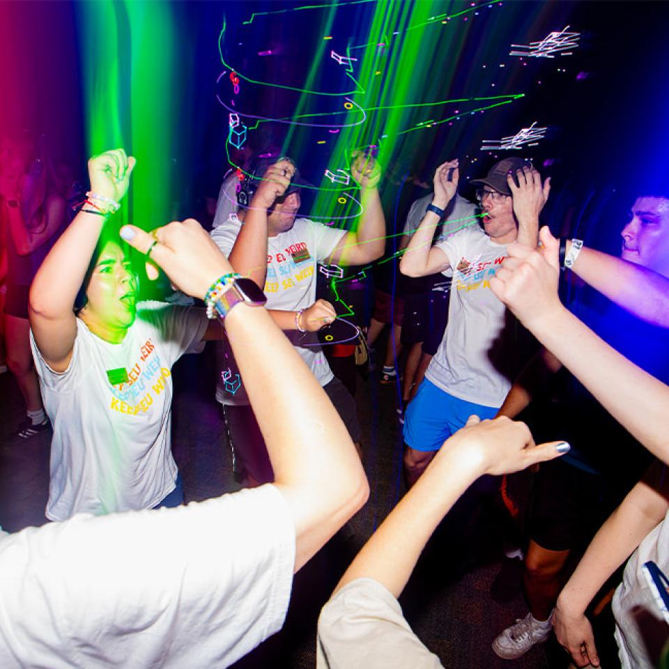 The image shows a group of young people dancing energetically at a silent disco. They are wearing headphones and colorful light beams illuminate the dark room, creating a vibrant atmosphere. Many are dressed in casual attire, including T-shirts that read "Keep SEU Weird." The dancers are raising their hands, clapping, and moving to the music, displaying expressions of joy and excitement. The lively and immersive environment highlights the fun and engaging nature of the event.