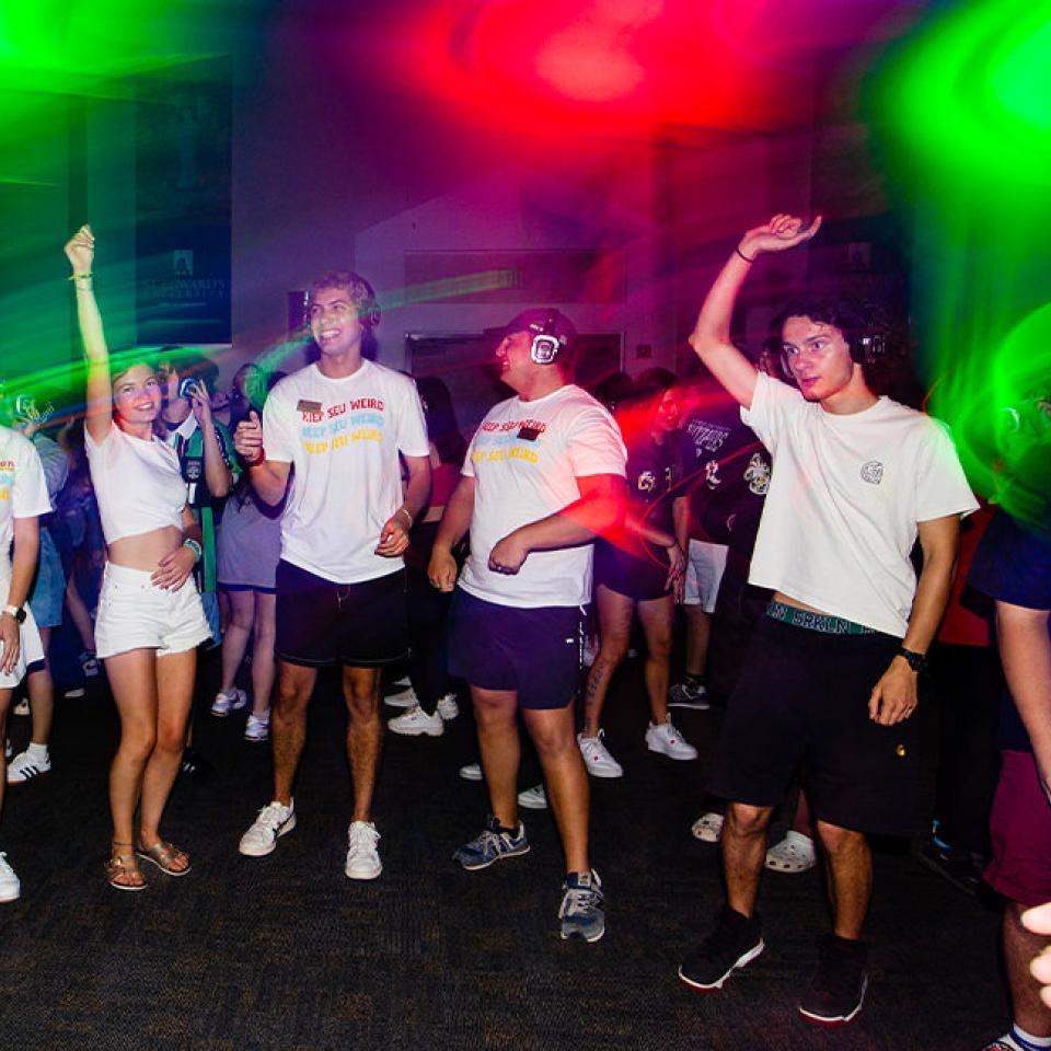 The image captures a group of young people dancing at a silent disco. They are all wearing headphones and are illuminated by colorful, vibrant lights. Many are dressed in casual attire, with some wearing T-shirts that say "Keep SEU Weird." The atmosphere is energetic and joyful, with participants smiling, raising their hands, and moving to the music. The dark room and the colorful light beams create a lively and immersive party scene.