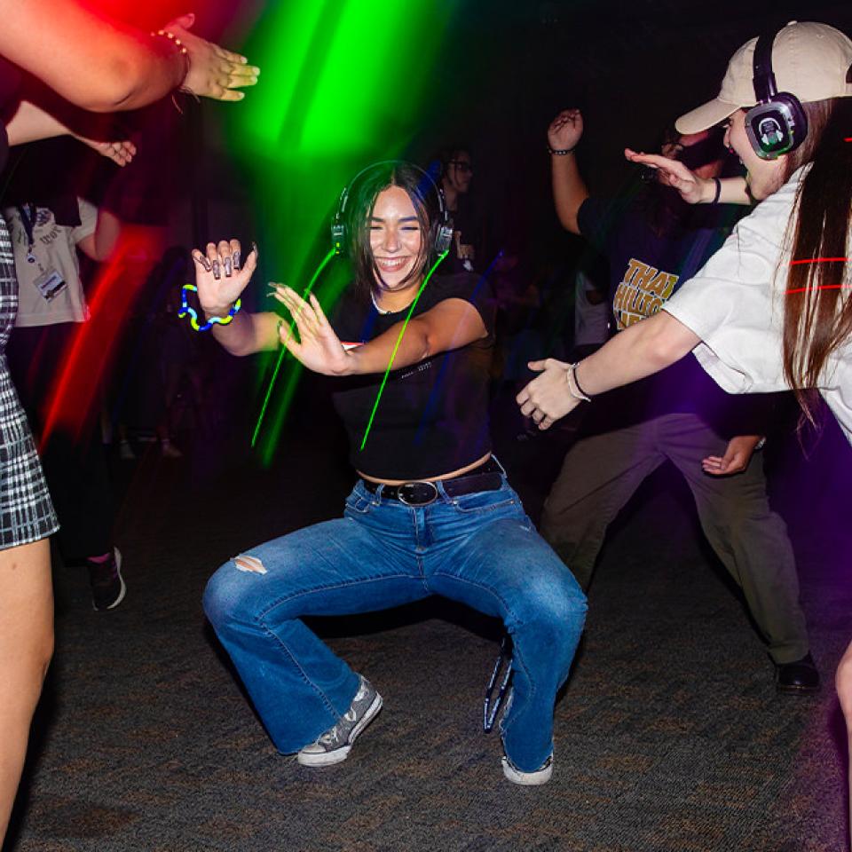 The image captures a lively scene at a silent disco. A young woman in a black top and ripped jeans is energetically dancing in the center, wearing headphones. Around her, other dancers, also wearing headphones, are enjoying the music and dancing. The room is dark, illuminated by colorful lights and beams, creating a vibrant atmosphere. Everyone looks happy and engaged, suggesting a fun and immersive experience. The casual attire and joyful expressions highlight the relaxed and festive mood of the event.