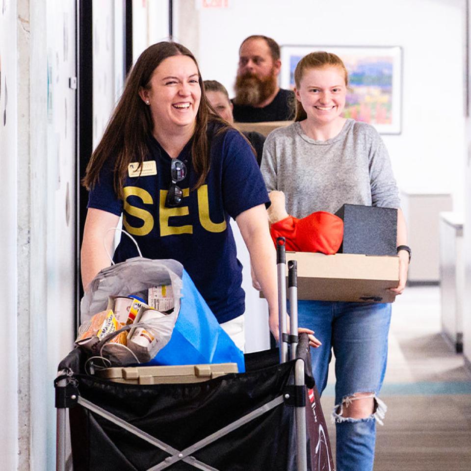 The image shows two smiling women moving belongings down a hallway. The woman in front is wearing a navy blue T-shirt with yellow letters "SEU" and is pushing a cart filled with various items, including a blue bag and snacks. The woman behind her is carrying a box and wearing a gray sweater and ripped jeans. There are two other people, a man and a woman, slightly out of focus in the background. The hallway has several doors and is brightly lit.