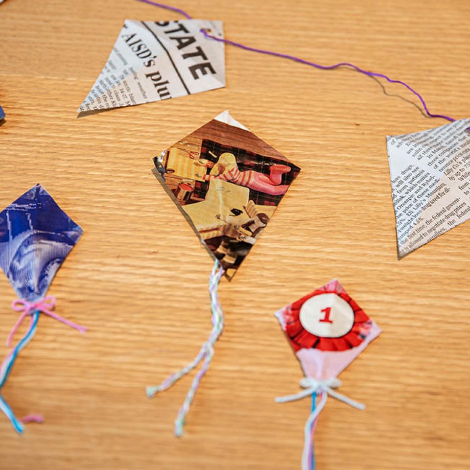 The image shows a table with small, handmade kites made from recycled materials such as magazine pages and newspapers. Each kite is decorated with colorful string tails tied in bows. The kites are arranged neatly on the table, showcasing their unique designs and patterns. This display is part of a craft project focused on upcycling, where old magazines and newspapers are creatively repurposed into decorative kite garlands.