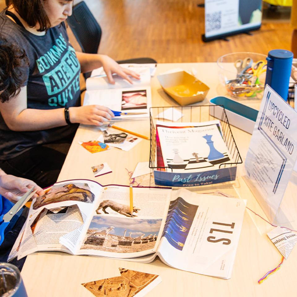 The image shows two individuals sitting at a table engaged in an arts and crafts activity. The person on the left, wearing a blue T-shirt, is cutting images from a magazine. The person on the right is also working with magazines and other craft materials. The table is scattered with various supplies, including scissors, colorful threads, and containers. Signs on the table read "Upcycled Kite Garland" and "Past Issues Free to Take," indicating an upcycling craft project.