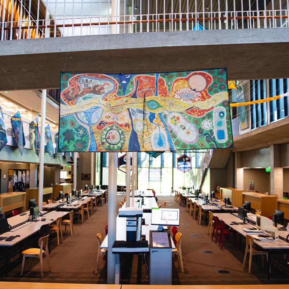 The image shows the interior of a large, modern building, likely a library or academic center, with colorful kites hanging from the ceiling. The kites display various artistic designs and are suspended above rows of desks and computer workstations. The architecture includes concrete beams, open spaces, and large windows that let in natural light. The setting is organized and bright, indicating a creative and vibrant atmosphere, possibly part of an exhibition or cultural display.