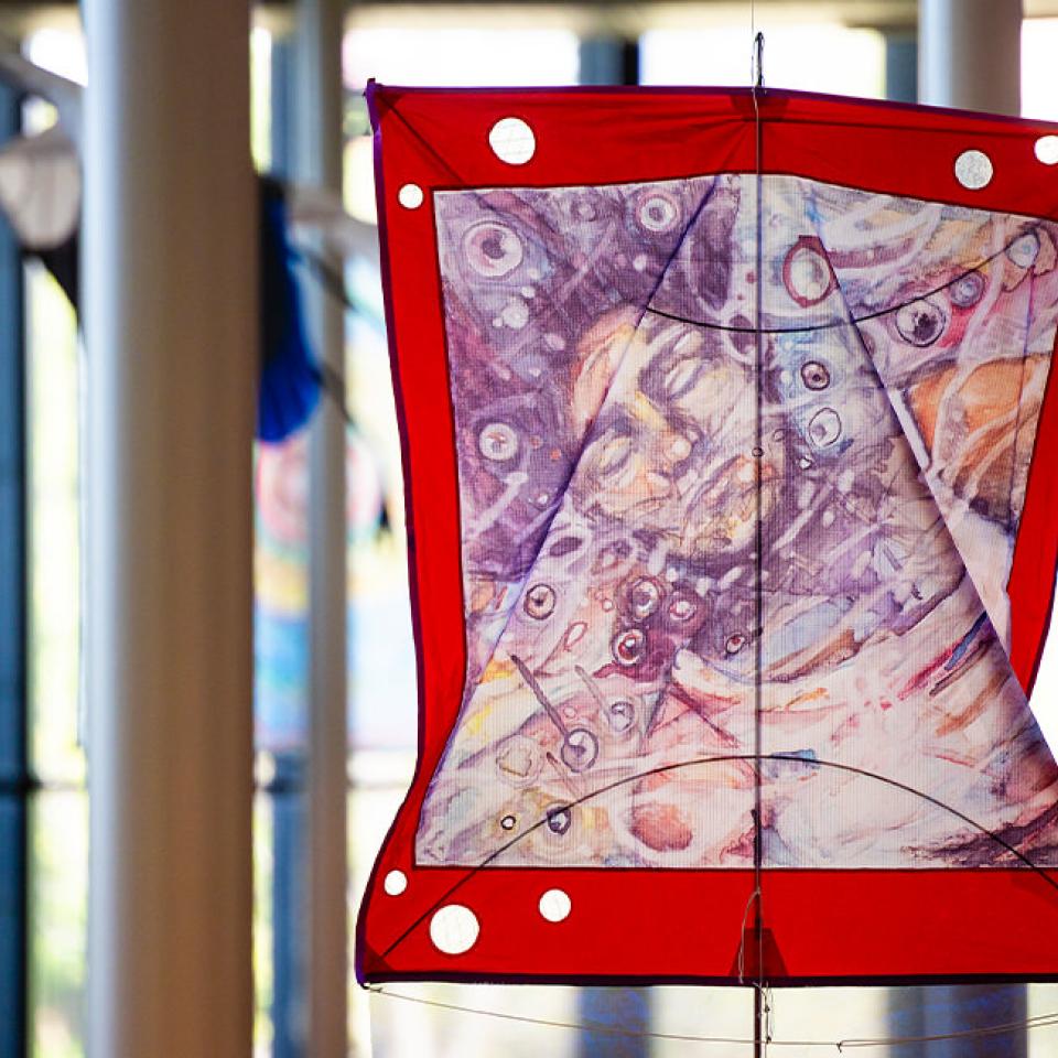 The image shows a colorful kite displayed indoors, hanging from the ceiling. The kite has a vibrant red border with white circles and an abstract design in the center featuring swirling patterns and various shades of purple, pink, and beige. The background includes large windows and pillars, suggesting the kite is part of an exhibition in a spacious, well-lit building. The setting is artistic and modern, with other kites visible in the distance.