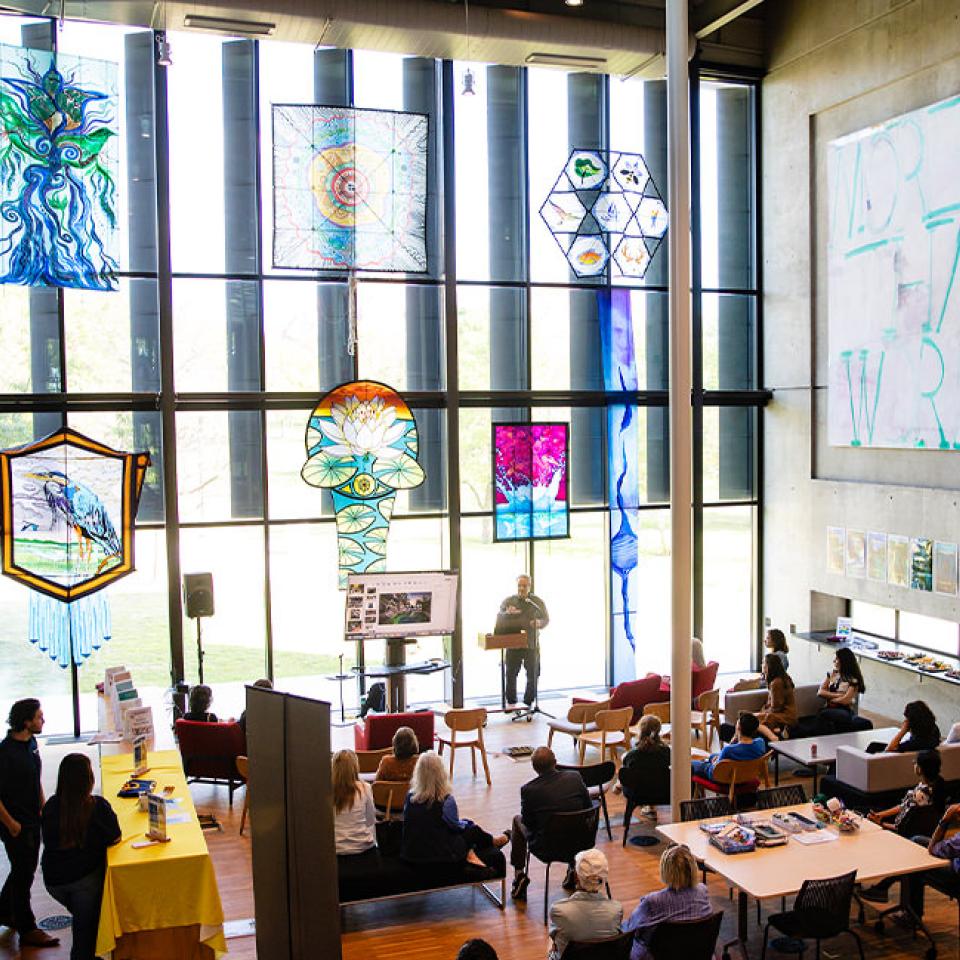 The image shows a large, modern indoor space with floor-to-ceiling windows and high ceilings, showcasing colorful kites hanging from the ceiling. The kites have various artistic designs, including abstract patterns and nature scenes. Below, people are seated and standing, attending a presentation or event. A speaker is at a podium near a screen displaying images. The setting includes desks, chairs, and a vibrant, open atmosphere, suggesting a creative or educational event.