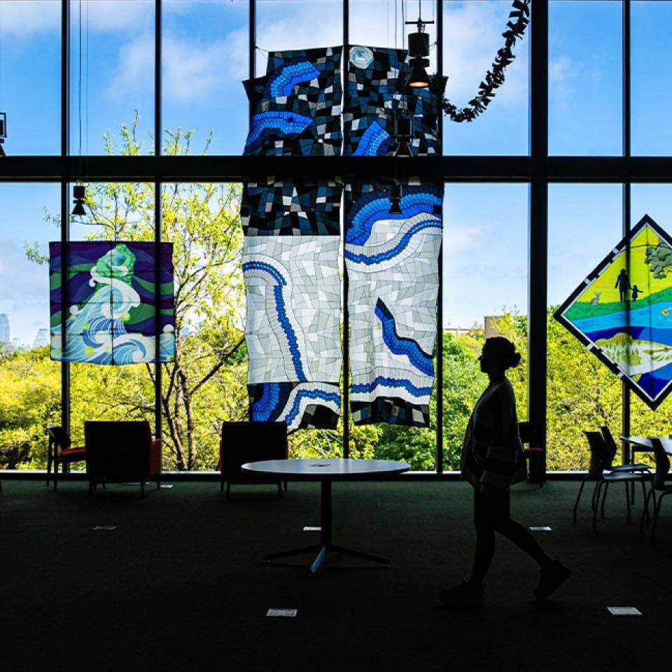 The image shows a large room with floor-to-ceiling windows displaying colorful kites hanging in front of the glass. The kites feature various designs, including abstract patterns and nature scenes. A person silhouetted against the light is walking through the room, adding a sense of scale. Outside, lush green trees and a clear blue sky are visible, suggesting a serene setting. The atmosphere is artistic and contemplative, likely part of an exhibition or gallery.