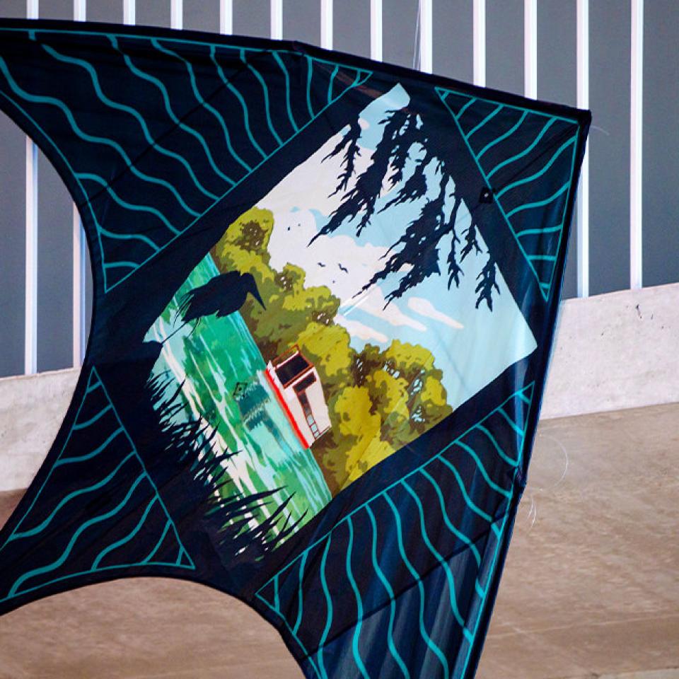 The image shows a kite displayed indoors, featuring a scenic design of a boat on a green river surrounded by trees and a bird perched on the bank. The kite is shaped like a delta and has a dark border with wavy blue lines. The background includes a concrete surface and vertical metal bars, indicating it is part of an exhibition or display. The artwork on the kite is detailed and vibrant, capturing a tranquil, nature-inspired scene.