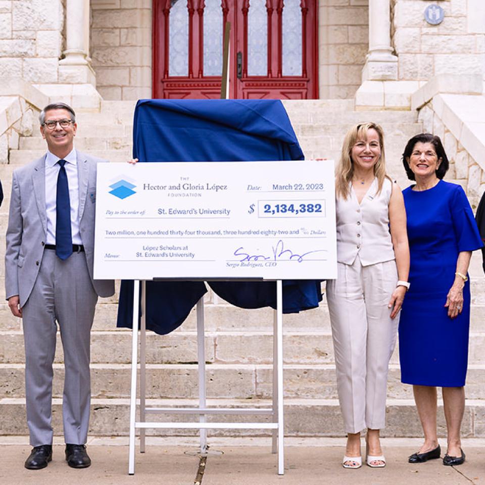 The image shows five individuals standing in front of a stone building, holding a large ceremonial check from the Hector and Gloria López Foundation made out to St. Edward's University for $2,134,382. The group includes a young woman in a navy top and white pants, a man in a light gray suit, a woman in a white outfit, another woman in a blue dress, and a man in clerical attire. They are all smiling, indicating a celebratory occasion, likely related to a significant donation or grant announcement.