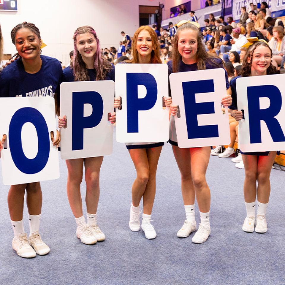 The image shows a group of seven women standing in a gymnasium, holding large signs that spell out "TOPPERS." They are all smiling and wearing matching "St. Edward's Hilltoppers" T-shirts and athletic shoes. The setting suggests a pep rally or school spirit event, with a crowd of people seated in the background, cheering and participating in the event. The atmosphere is energetic and celebratory.