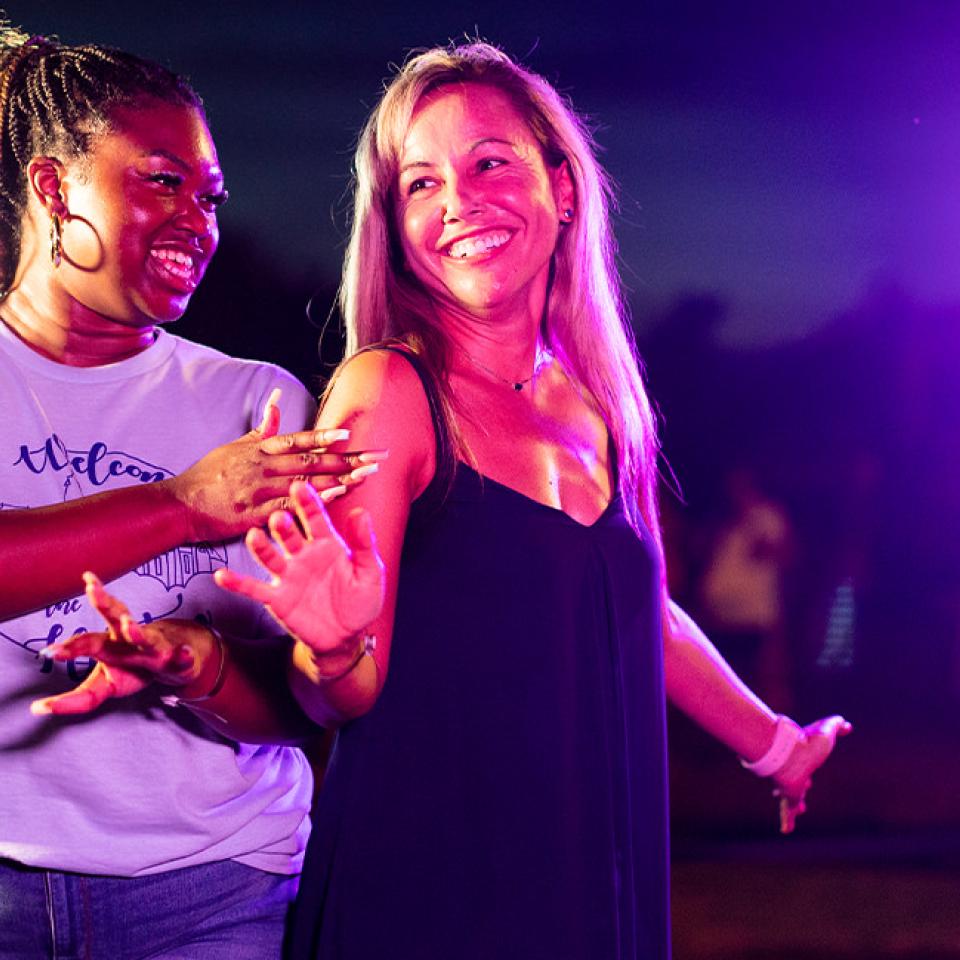 The image shows two women dancing and smiling under vibrant purple lighting at an outdoor event. The woman on the left is wearing a light-colored T-shirt with text, while the woman on the right is in a dark sleeveless top. They are enjoying themselves, with the woman on the left playfully guiding the woman on the right. The background is blurred, indicating a nighttime setting with other people and festive activities in the vicinity.