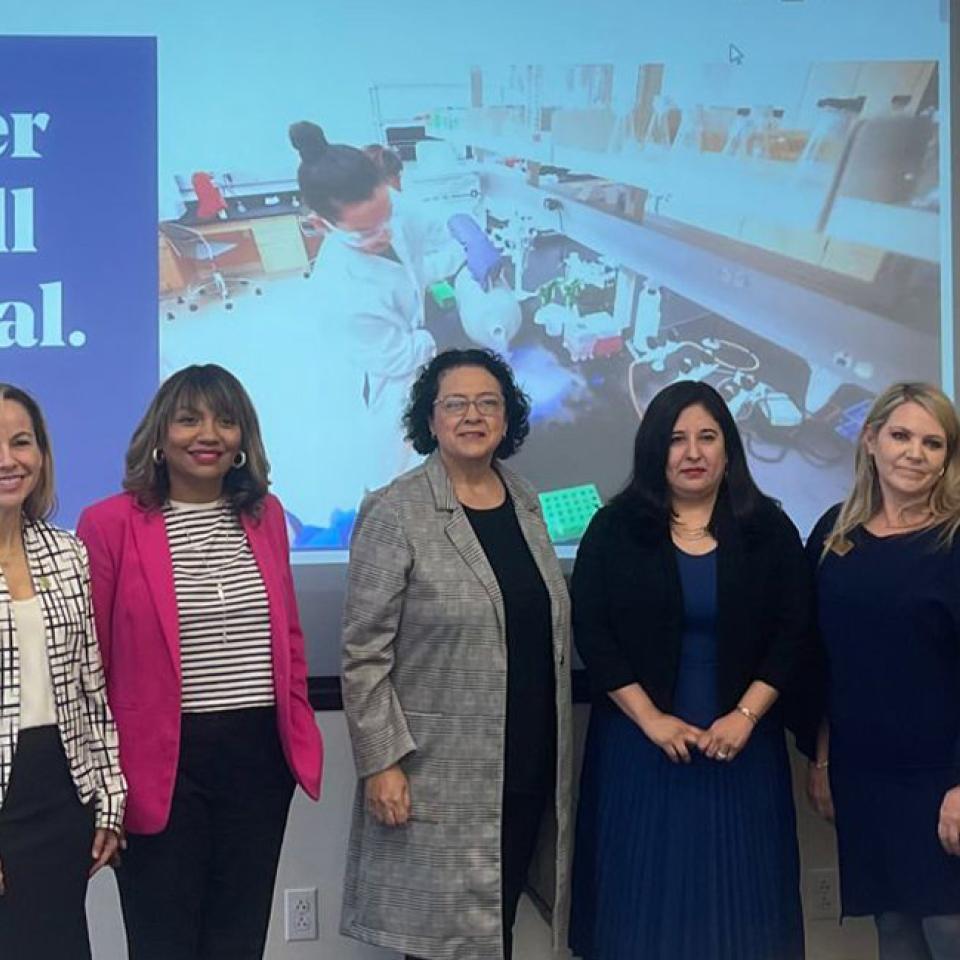 The image shows six women standing in front of a projected presentation slide that reads "Discover your full potential" and features an image of a lab setting. They are dressed in professional attire, including blazers and dresses, and are smiling at the camera. The setting suggests they are participating in a professional or educational event, possibly a roundtable discussion or a conference related to business or academic achievement. The atmosphere is positive and empowering.