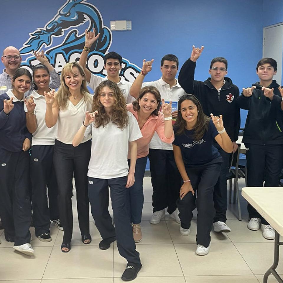 The image shows a group of students and adults standing in a classroom at Balboa Academy, making the "Toppers Up" hand gesture and smiling at the camera. The group consists of both males and females, dressed in school uniforms and casual attire. They are standing in front of a blue wall with the school's logo, indicating a school-related event or visit. The atmosphere is cheerful and enthusiastic, suggesting a positive and engaging occasion.