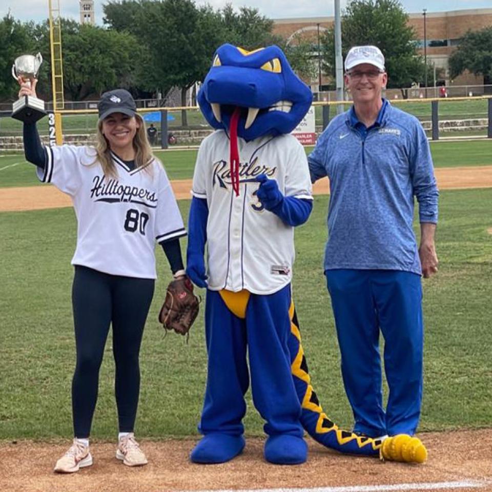 The image shows three individuals standing on a baseball field. On the left, a woman in a "Hilltoppers" jersey holds up a trophy and a baseball glove, smiling. In the middle, there is a person in a blue and yellow mascot costume with "Rattlers" on the jersey. On the right, a man in a blue athletic outfit and cap stands smiling. The background features a baseball diamond and some buildings, indicating a sports event, possibly a friendly competition or a ceremonial presentation.