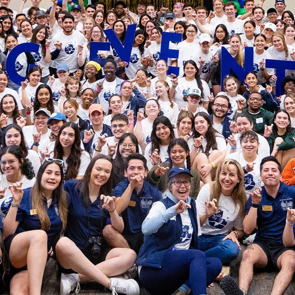 The image shows a large group of enthusiastic people posing together for a group photo. They are dressed in matching T-shirts and appear to be part of a community or volunteer event, as indicated by the large, blue letters held up by some participants spelling "BIG EVENT." The group is diverse, with many individuals smiling and making hand gestures. The atmosphere is lively and celebratory, suggesting a successful and enjoyable event.