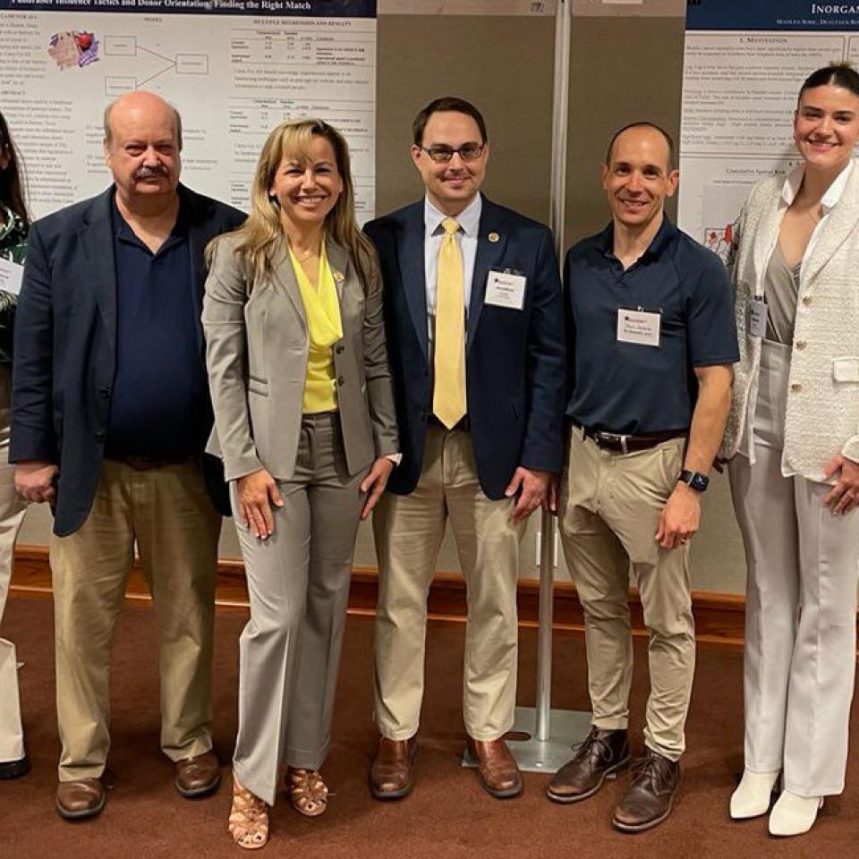 The image shows six individuals standing in front of research posters, likely at an academic or scientific conference. They are dressed in business or business casual attire, including blazers, shirts, and trousers. The group consists of three women and three men, all smiling at the camera. The setting suggests they are presenting or discussing research findings, indicated by the detailed posters displayed behind them. The atmosphere is professional and collegial.